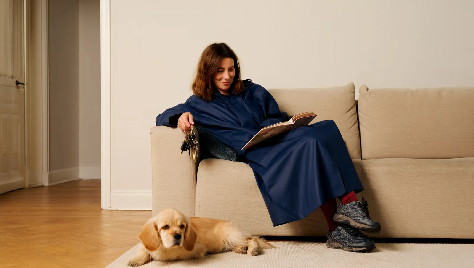 A woman in a navy raincoat and outdoor trainers sitting reading on a sofa with a bunch of house keys dangling from one hand while a golden puppy dozes on the rug beside her