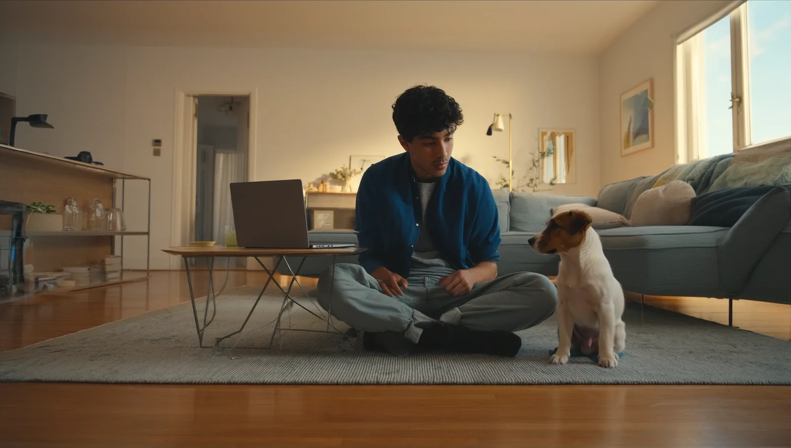 A man sitting on a living room floor with an open laptop and an attentive puppy beside him during an at-home training session.